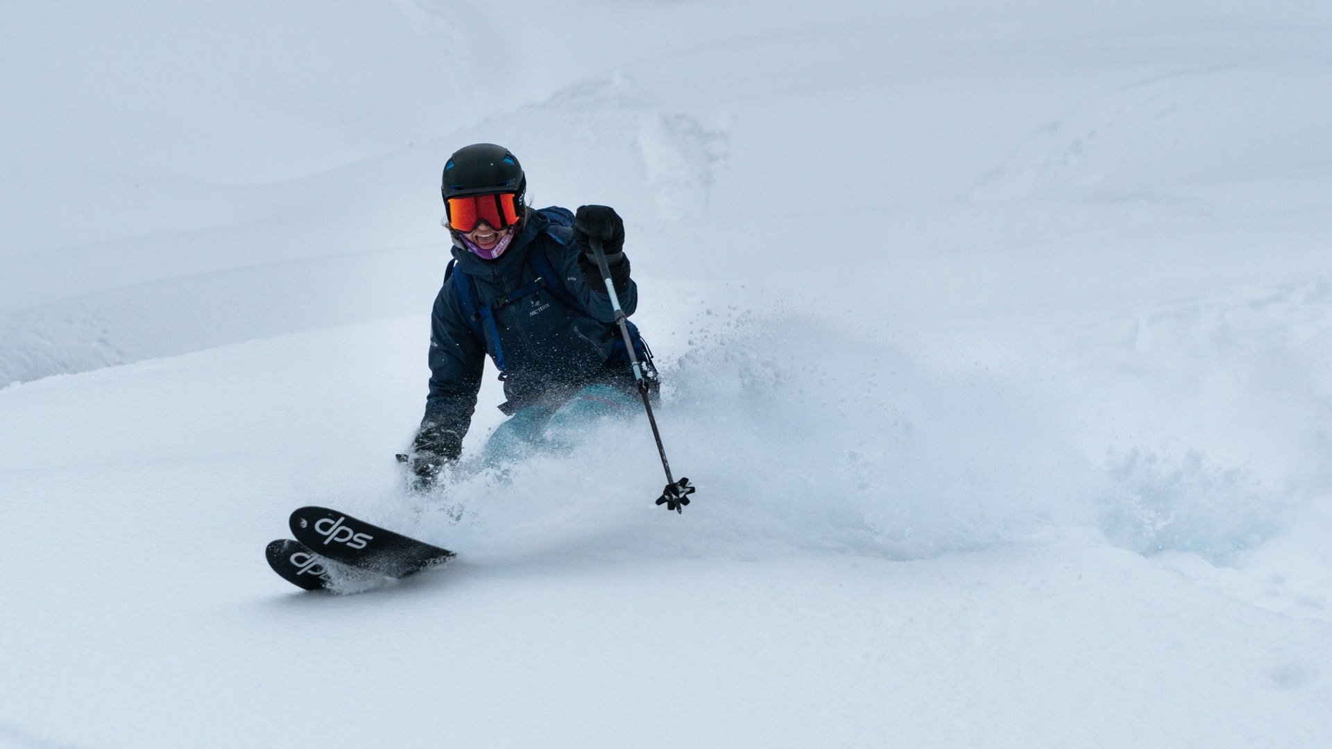 skier in blue jacket and orange googles slashing a turn in powder